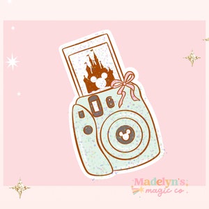 May include: A light blue and white glitter camera with a pink bow. The camera has a photo inside of a brown silhouette of Cinderella's castle with Mickey Mouse ears on top. The text "Madelyn's magic co." is at the bottom of the image.