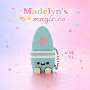 May include: A light blue surfboard-shaped keychain with a pink hibiscus flower graphic, a smiling face, and two small white feet. The text Madelyn's magic co is at the top, and the text click click is on the right side with a metal ball chain.
