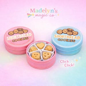 May include: Pink and blue round tins of shortbread cookies with smiling cookie illustrations. The pink tin is open, revealing various cookie shapes. The text "Shortbread Cookies" is printed on the tins. The image also includes the text "Madelyn's magic co."