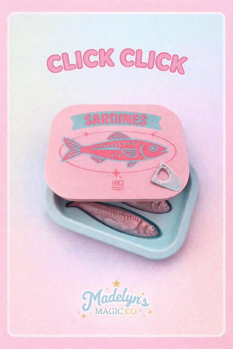May include: A pink sardine can with a teal label that reads "Sardines" and a red fish illustration. The can is open, revealing two sardines in a light blue tray. The text "CLICK CLICK" is at the top, and "Madelyn's Magic Co." is at the bottom.
