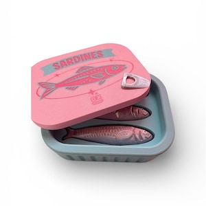 May include: A pink sardine can with a blue interior and lid. The lid features a red fish illustration and the word "Sardines" in blue. Two sardines are visible inside the can.