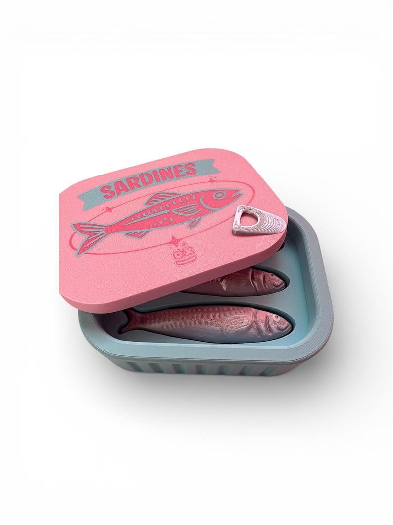 May include: A pink sardine can with a blue interior and lid. The lid features a red fish illustration and the word "Sardines" in blue. Two sardines are visible inside the can.