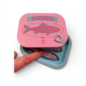 May include: A pink sardine tin with a teal and pink fish illustration and the word "SARDINES". The tin is open, revealing two sardines in a teal container. A finger touches one of the sardines.