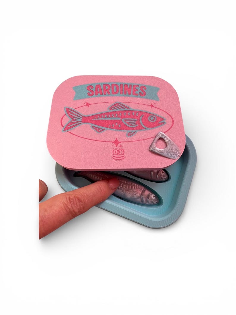 May include: A pink sardine tin with a teal and pink fish illustration and the word "SARDINES". The tin is open, revealing two sardines in a teal container. A finger touches one of the sardines.