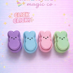 May include: Four pastel-colored bunny-shaped objects in purple, blue, pink, and green, each with black dot eyes. The image has text that says "Madelyn's magic co" and "CLICK CLICK!". The background is a soft gradient of pink and blue.