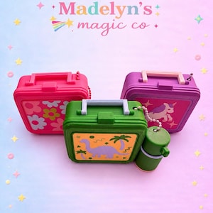 May include: Three miniature plastic lunch boxes with handles and ball chains. One is pink with a floral pattern, one is purple with a unicorn graphic, and one is green with a dinosaur and palm tree design, accompanied by a matching green thermos.