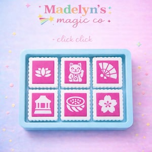 May include: A light blue tray holding six pink and white stamps with various designs, including a lotus flower, a cat, and a fan. The text "Madelyn's magic co" and "click click" are at the top.