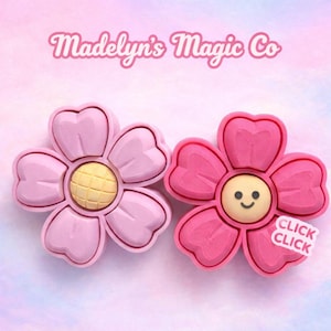 May include: Two pink flower-shaped accessories with yellow centers and a smiling face. One flower is a lighter pink, the other is a darker pink. The text "Madelyn's Magic Co" is at the top. The words "Click Click" are on a sticker on one flower.
