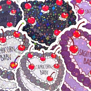 May include: A selection of holographic stickers featuring various zodiac signs with the text "Baby" on each sticker. The stickers are shaped like heart-shaped cakes with cherries on top. The stickers are displayed against a white background.