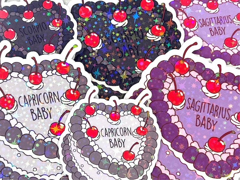 May include: A selection of holographic stickers featuring various zodiac signs with the text "Baby" on each sticker. The stickers are shaped like heart-shaped cakes with cherries on top. The stickers are displayed against a white background.