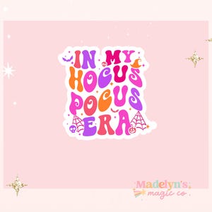 May include: A sticker with the text "IN MY HOCUS POCUS ERA" in a retro font, with Halloween-themed illustrations. The sticker is on a pink background with gold star accents and the text "Madelyn's magic co."