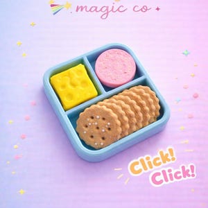 May include: A light blue divided container filled with a yellow cheese square, a pink circle, and several round crackers. The image has the text "Madelyn's magic co." and "Click! Click!" on a pastel purple and pink background.