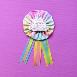 May include: A colorful fabric rosette with a rainbow tie, featuring an embroidered cake with the text "It's My Birthday".