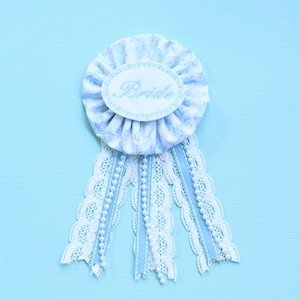 May include: A blue and white fabric rosette with the word "Bride" embroidered on it. The rosette is decorated with white lace and blue ribbon.