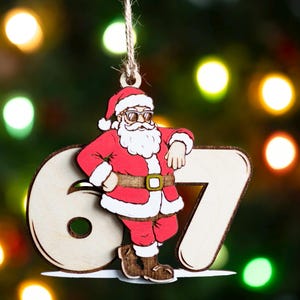May include: Wooden Christmas ornament depicting Santa Claus in sunglasses, leaning against the numbers "67". Santa is dressed in a red suit with white trim and a brown belt. The ornament hangs from a twine string against a blurred background of Christmas lights.