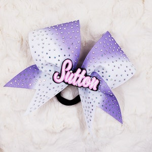 Op de afbeelding: Paarse en witte cheer bow met de naam "Sutton" in roze glitter in het midden.