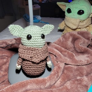 K&ouml;nnte beinhalten: Zwei geh&auml;kelte Baby Yoda Pl&uuml;schtiere in Gr&uuml;n und Braun auf einer braunen flauschigen Decke. Ein Pl&uuml;schtier tr&auml;gt einen braunen geh&auml;kelten Mantel.