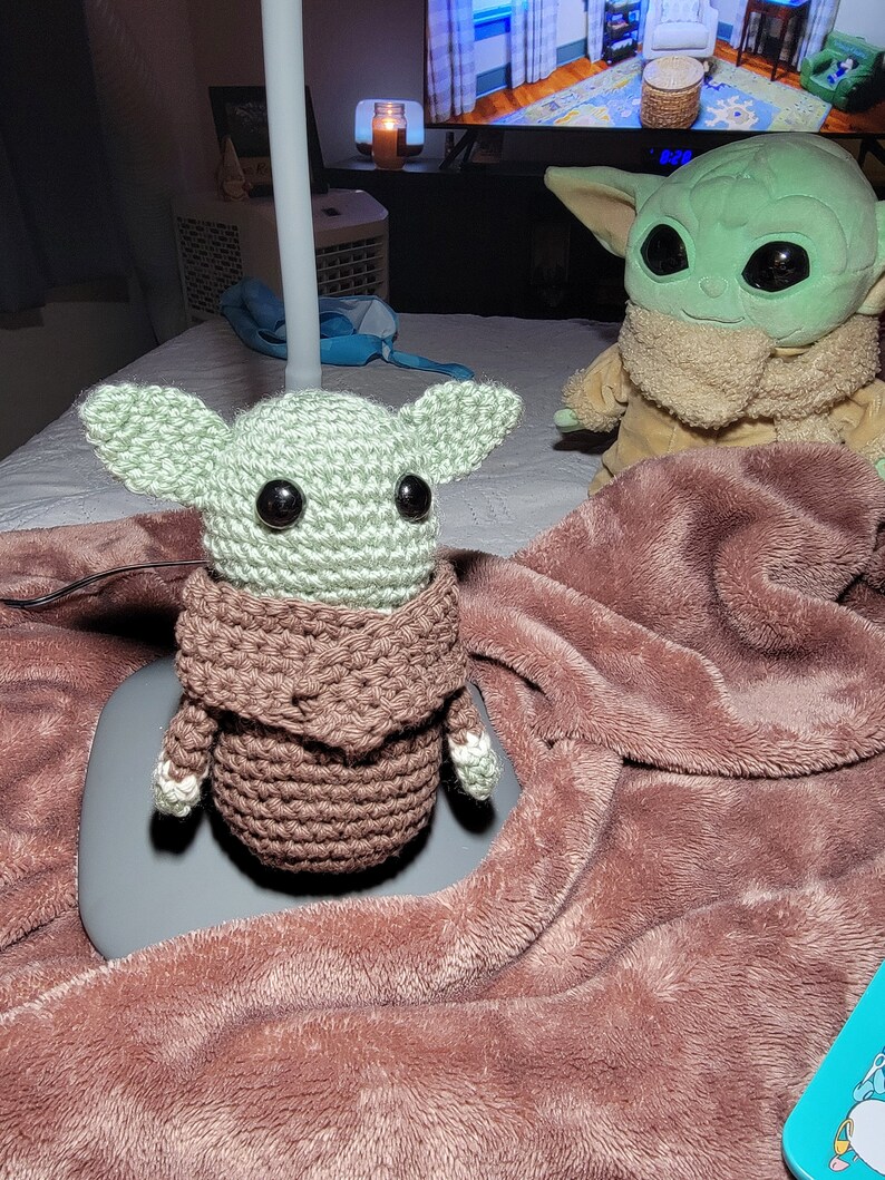 K&ouml;nnte beinhalten: Zwei geh&auml;kelte Baby Yoda Pl&uuml;schtiere in Gr&uuml;n und Braun auf einer braunen flauschigen Decke. Ein Pl&uuml;schtier tr&auml;gt einen braunen geh&auml;kelten Mantel.