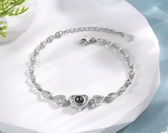 May include: A silver bracelet with a heart-shaped charm that projects a message when viewed from a certain angle. The bracelet is made of a delicate chain with small heart links. The charm is adorned with small clear stones.
