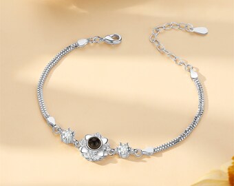 May include: A silver chain bracelet with a small flower charm that projects a message when viewed with a light source. The charm is surrounded by small clear stones.