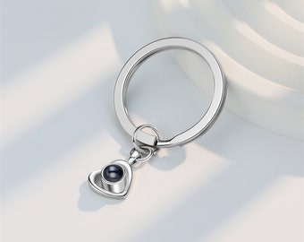May include: Silver heart-shaped keychain with a small black circle in the center. The keychain has a silver ring attached to it.
