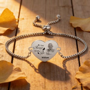 Op de afbeelding: Zilverkleurige armband met een hartvormige hanger. De hanger heeft een foto van een persoon, de woorden "I Am Always With You" en de naam "William". De armband ligt op een houten oppervlak met gele bladeren.