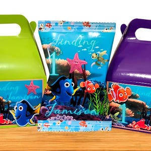 Peut inclure: Une boîte à poignée vert citron, une boîte à poignée violette et un sachet de friandises central, ornés de graphismes des personnages Nemo et Dory, d'un récif corallien, du texte Finding Jamison et du chiffre 1. Une étiquette logo indique Everly Hope Creations.