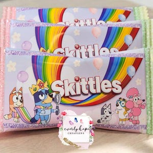 Könnte beinhalten: Drei rechteckige Skittles-Süßigkeitenverpackungen mit hellviolettem Hintergrund, Regenbogen, Luftballons und Zeichentrick-Hunden. Die Verpackung zeigt einen Hund mit Krone und Lupe sowie einen rosa Pudel. Ein Logo trägt die Aufschrift Everly Hope Creations.