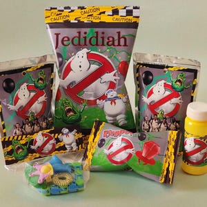 Könnte beinhalten: Eine Sammlung von Party-Mitbringseln und Leckereien im Ghostbusters-Stil. Enthält Süßigkeitenbeutel, Ring Pops und eine Seifenblasenflasche, alle mit dem ikonischen Logo und den Charakteren. Die Verpackung hat ein Warnband-Design.