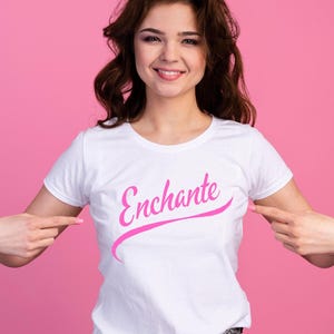 Pode incluir: Camiseta branca com a palavra "Enchante" em escrita rosa. A camiseta está sendo apontada por uma pessoa. O fundo é de cor rosa sólida.