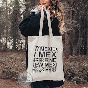 May include: A beige tote bag with black text that reads "W MEXICO / MEXI NEW MEXICO NEW MEXICO NE NEW MEXI V MEXICO NEW MEXICO NEW MEXICO NEW MEXICC IE)"  The bag is being held by a person in a forest.