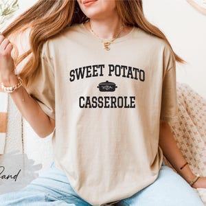 May include: A sand-colored t-shirt with the words "SWEET POTATO CASSEROLE" printed in black, along with a small pot illustration. The shirt is a casual, comfortable style, perfect for those who love cooking or enjoy comfort food.