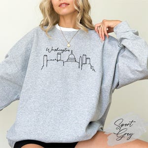 May include: A light gray sweatshirt with a white crew neck collar. The front features a black line drawing of the Washington, D.C. skyline with the word "Washington" above it. The lower right corner has the words "Sport Grey".