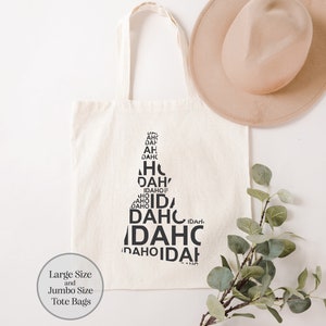May include: A white canvas tote bag with a black graphic design of the word "Idaho" repeated in different orientations. The text is arranged to form the shape of the state of Idaho. The bag is available in large and jumbo sizes.