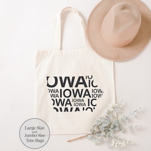 May include: A white canvas tote bag with the word "IOWA" repeated in a circular pattern in black text. The bag is on a white surface with a brown hat and a sprig of greenery.