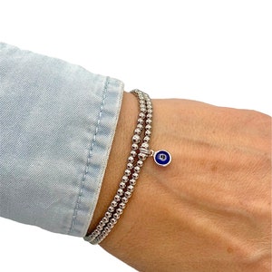 May include: A silver beaded bracelet with a small blue and white evil eye charm. The bracelet is made of small, round beads and is worn on a wrist. The charm is a small, round pendant with a blue eye design.