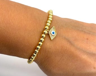 May include: Gold beaded bracelet with a diamond-shaped charm. The charm features a blue eye design with white and clear accents. The bracelet is on a wrist against a white background.