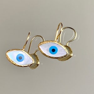 May include: A pair of gold-colored earrings featuring an eye design. The earrings have a white background with a blue iris and a black pupil. The gold-colored frame has a textured appearance.