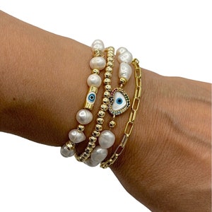May include: Four gold bracelets are displayed. One is a chain, the others feature white pearls and gold beads. Two bracelets have an evil eye charm, one with a blue eye and the other with a heart-shaped design.