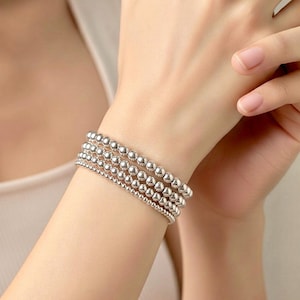 May include: Five silver beaded bracelets of different sizes are stacked on a wrist. The bracelets are made of small, round, shiny beads. The skin tone is light. The bracelets are a simple and elegant accessory.