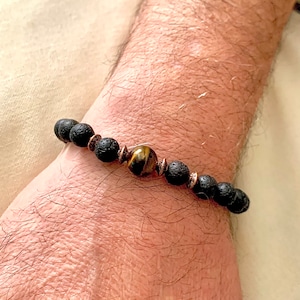 May include: A bracelet with black lava rock beads, a central tiger's eye bead, and copper-coloured spacers. The bracelet is displayed on a wrist, highlighting the natural textures and colours of the materials.