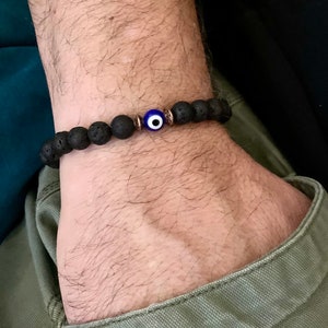 May include: Black lava rock bead bracelet with a blue and white evil eye charm. The bracelet is strung on a black elastic cord and features a bronze-colored spacer bead. The bracelet is worn on a wrist.