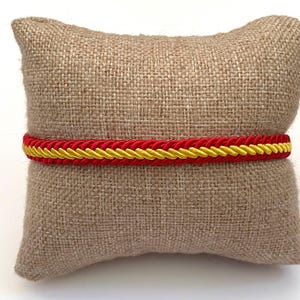 May include: A red and yellow braided bracelet, presented on a small, tan burlap cushion. The bracelet has a bright red outer braid and a yellow inner braid. The cushion's neutral tone accentuates the bracelet's colours and intricate design.