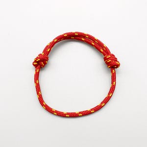 Op de afbeelding: Een rood en geel touw armband met verstelbare knopen. De armband is gemaakt van een duurzaam, geweven touwmateriaal. De knopen zijn geel en het touw is rood met gele accenten. De armband is ontworpen om om de pols te worden gedragen.