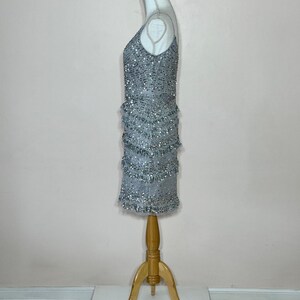 May include: A silver beaded mini dress with a V-neckline and fringe detailing. The dress is displayed on a mannequin with a wooden stand. The dress is sleeveless and has a fitted silhouette.