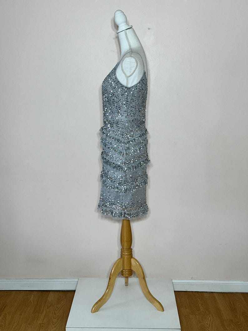 May include: A silver beaded mini dress with a V-neckline and fringe detailing. The dress is displayed on a mannequin with a wooden stand. The dress is sleeveless and has a fitted silhouette.