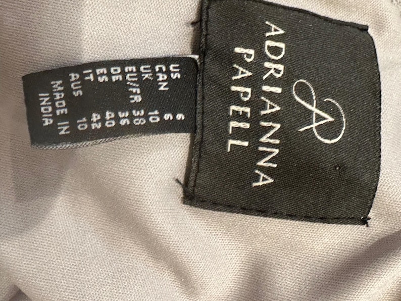 May include: A close-up of a black label with the brand name "ADRIANNA PAPELL" in white lettering. The label also includes size information for US, CAN, UK, EU/FR, DE, ES, IT, and AUS. The label reads "MADE IN INDIA".