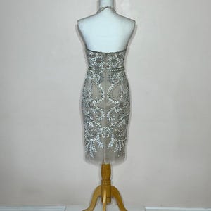Puede incluir: Vestido beige hasta la rodilla con intrincados bordados blancos y plateados. El vestido se exhibe en un maniqu&iacute; con un soporte de madera. El vestido de estilo halter presenta un corpi&ntilde;o ajustado y una falda transparente de tul.