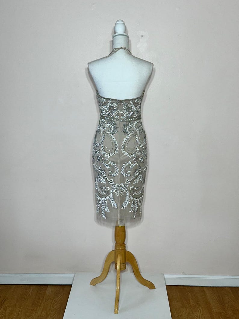 Puede incluir: Vestido beige hasta la rodilla con intrincados bordados blancos y plateados. El vestido se exhibe en un maniqu&iacute; con un soporte de madera. El vestido de estilo halter presenta un corpi&ntilde;o ajustado y una falda transparente de tul.