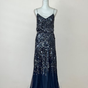 May include: A navy blue evening gown on a mannequin. The dress features a V-neckline, spaghetti straps, and is heavily embellished with sequins. The skirt flows into a sheer, dark blue hemline, creating a glamorous silhouette.